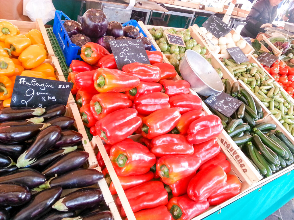 The Marche aux Fleurs in Nice, a fruit, vegetable, and artisanal foods market in Old Town. In the photo are various vegetables, including red peppers, eggplant, zucchini, nad tomatoes. 