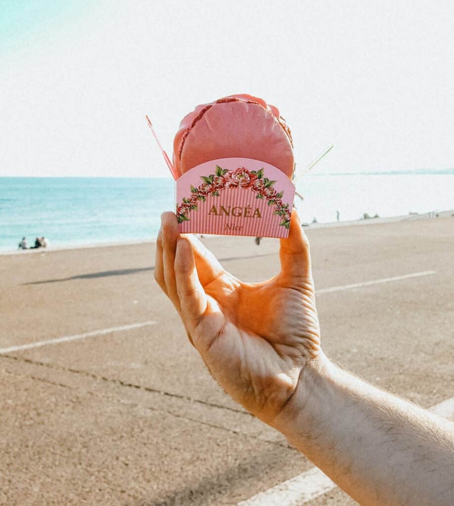A fancy macaron-gelato sandwich is held up in front of the Promenade des Anglais and the Mediterranean Sea in Nice, France.