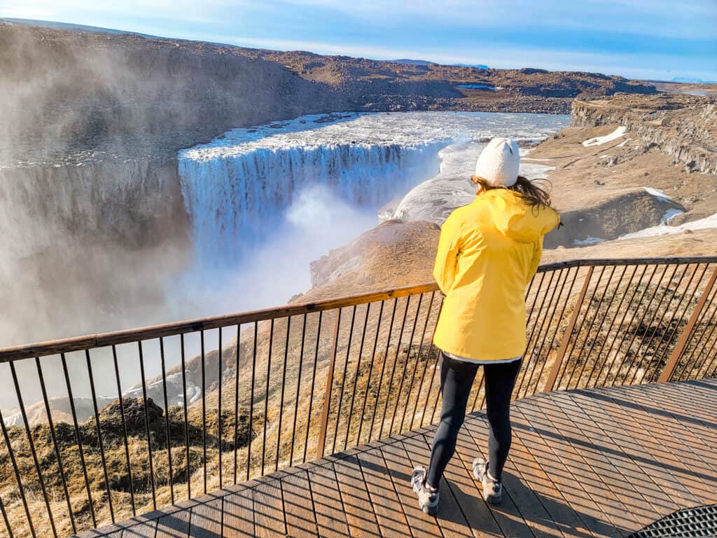 A woman in a yellow jackets looks at Dettifoss which is 150 ft tall and is one of the most powerfall waterfalls in all of Europe and is so white. 