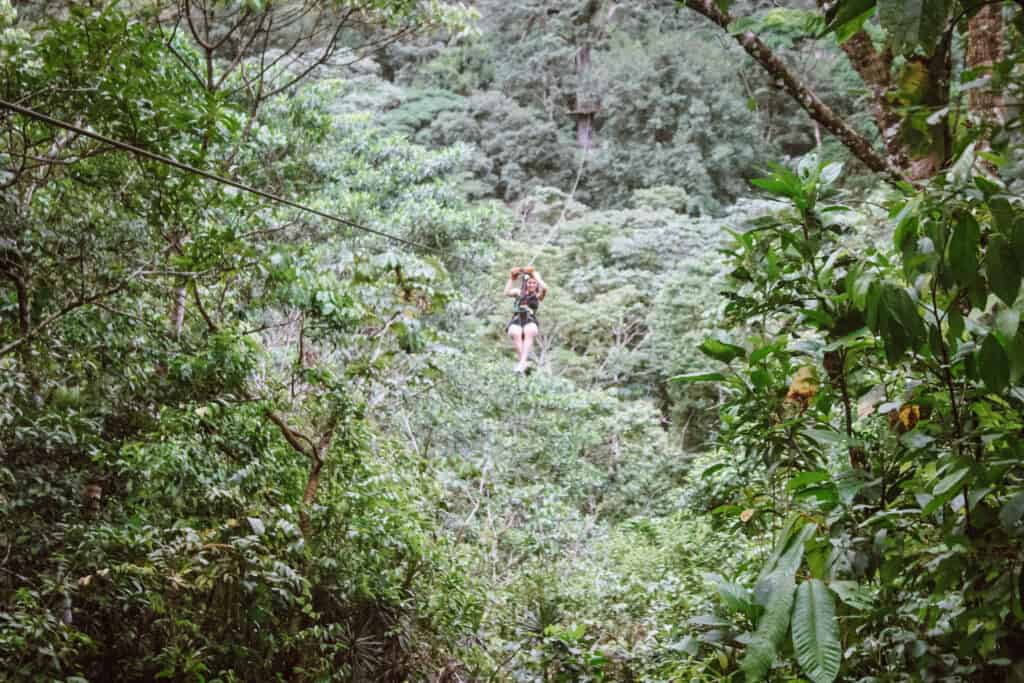 A woman ziplines through the rainforest of Costa Rica. All around her are trees and ferns. She is holding onto the zipline and coming towards the camera. 