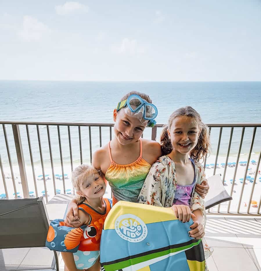 Three children in swimsuits, with arm floaties, goggles, and a boogie board are smiling at the camera. They are standing on a deck and you can see the beach and the blue waters of the Gulf of Mexico behind them. 