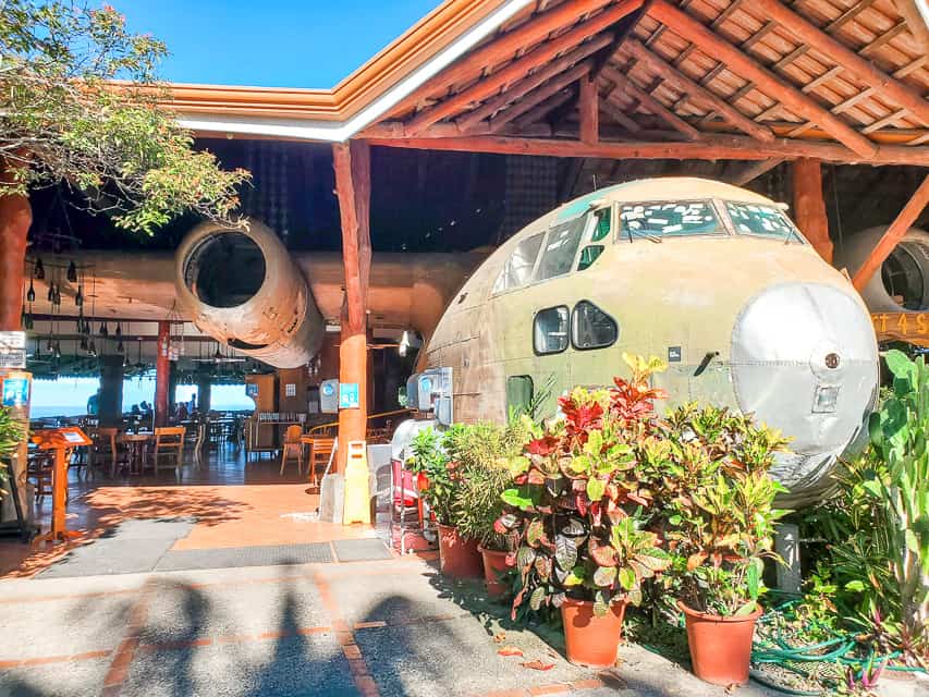 A restaurant is built around an old airplane. Potted plants decorate the sides of the airplane and you can see tables under the patio. The ocean is in the distance. 