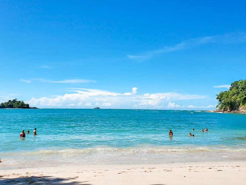 A strip of sandy beach is in the foreground, with beautiful blue ocean in the back. The skies are blue. There is some land and trees off to the sides. About 10 people are in the water swimming. 