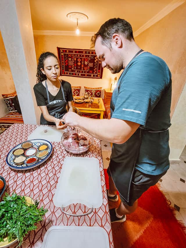 A Moroccan woman helps an American man measure spices to add to a bowl of  lamb meat. They are both wearing black aprons. The table they are standing at has a red and white tablecloth, white cutting boards, and herbs and a plate of spices are visible. A colorfully red seating area is in the background. 