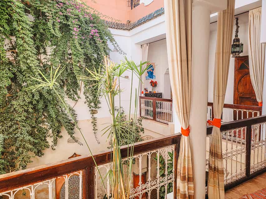 The view from a peaceful riad. A hallway and railing overlook a courtyard, with trees and plants growing in it. Curtains are tied back around the railing.