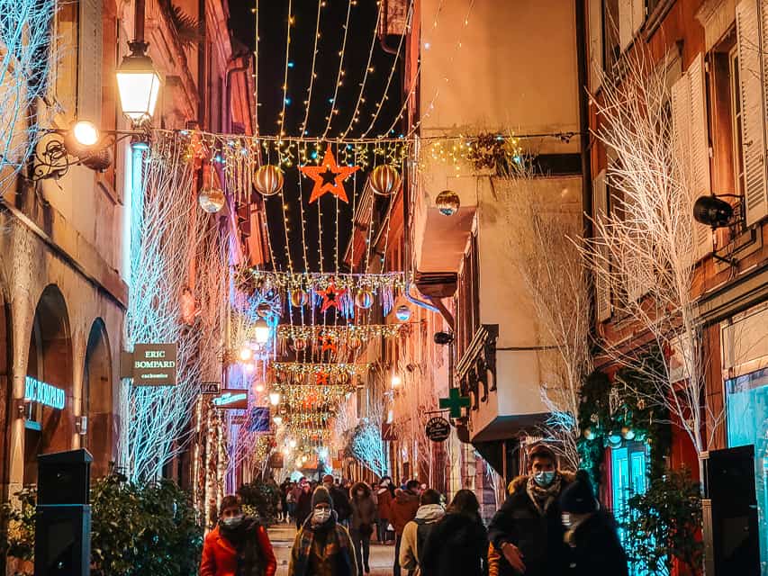 A Strasbourg street is illuminated by Christmas lights and decorations such as stars, golden ornaments, and lit trees. Rue des Orfevre.