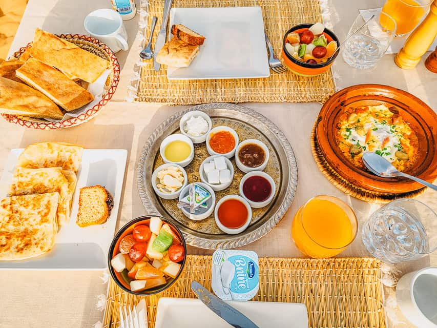 A breakfast offering in consisting of egg tagine, various jams and honeys, crepe, breakfast breads, fruit, yogurt, and juice.