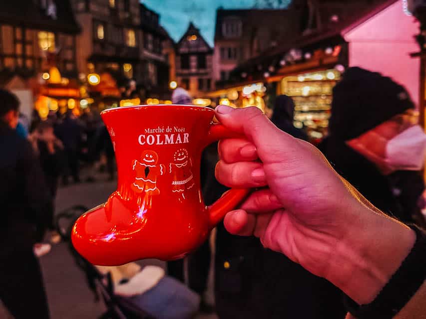 A hand holds a red, boot-shaped mug with the words, "Marche de Noel Colmar" and images of a gingerbread man and woman. A night-time Christmas market is in the background.
