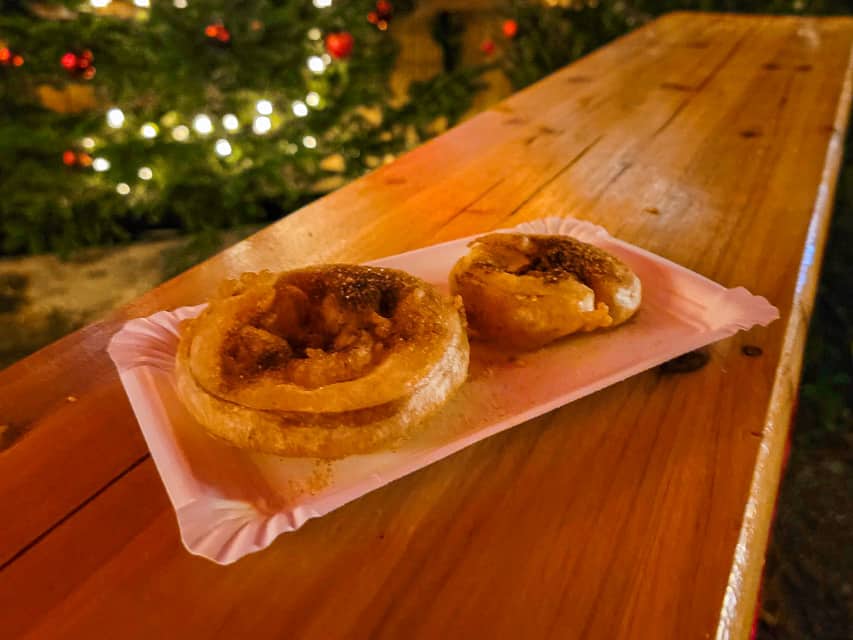 A white paper tray on a wooden table holds 2 apple fritters - fresh apple slices that were dipped in batter, fried, and covered in cinnamon. 