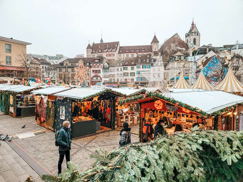 Overlooking the stalls of the Basel Christmas market, with big lit up stars on top of the stalls, and the buildings of Basel in the background. 