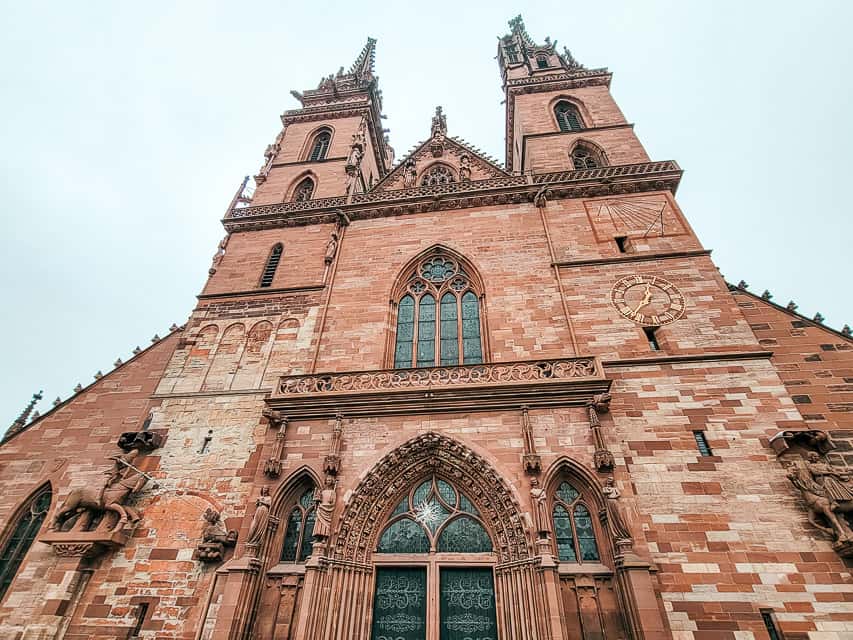 A view from the base of the Minster Basel Cathedral, loooking up to the spires. The cathedral is made of red brick and has two pointed spires. 