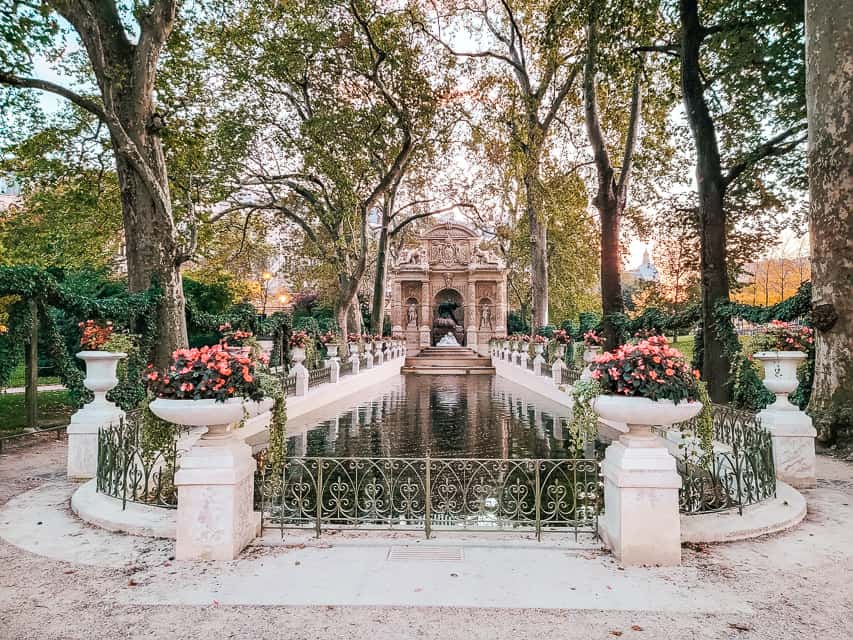 The Medici Fountain in the Jardin de Tuileries has a long basin of water lined by an iron railing and white flower pots, with a large sculpture piece behind it. 