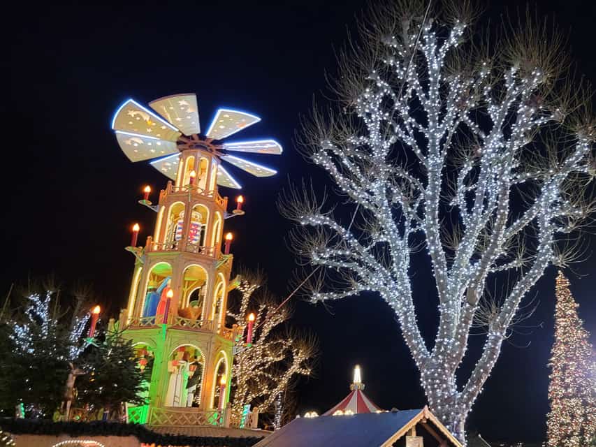 The top of a Christmas pyramid is in the foreground, with it's open towers with figures inside, and fan blades at the top of the tower. A tree lit with white lights is behind. 