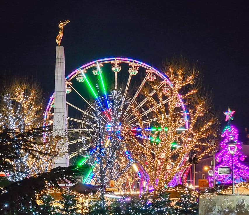 One of the main markets in Luxembourg at the Place de la Constitution, with a tall pillar, grand ferris wheel, games, and trees lit beautifully with thousands of lights. 