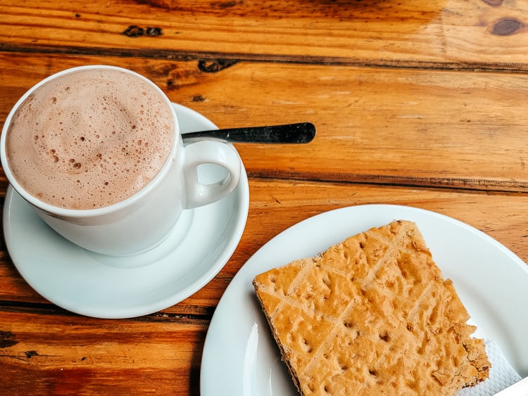 A cup of frothy hot chocolate - a traditional Colombian breakfast drink - and a square pastry to the side.