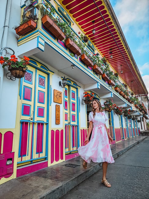 A woman in a pink dress steps off the curb on Calle Real, with a building decked out in red, blue, and yellow colors, with lots of flower pots and planter boxes around. 
