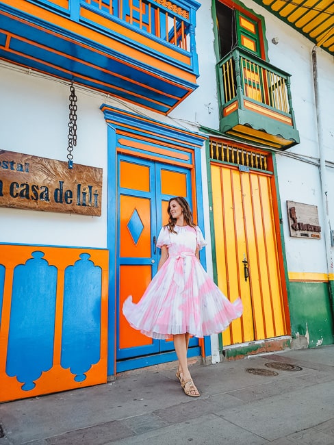 A woman in a pink and white dress stands smiling in front of a door with orange and blue, with a door in yellow, green, and orange next to her. 