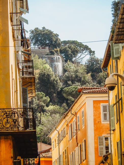 The waterfall on Castle Hill is visible in the greenery above Old Town in Nice. Golden buildings frame in the waterfall. 