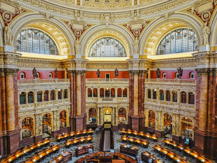 The reading room in the library of congress is semicircular with arches, columns, red walls, and numerous tables.
