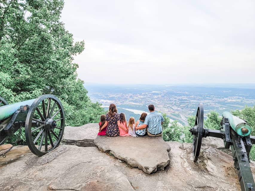 A family that includes 4 little girls sits on rocks at the edge of a mountain, with two historic cannons on either side, and views over the Tennessee River and Chattanooga. 