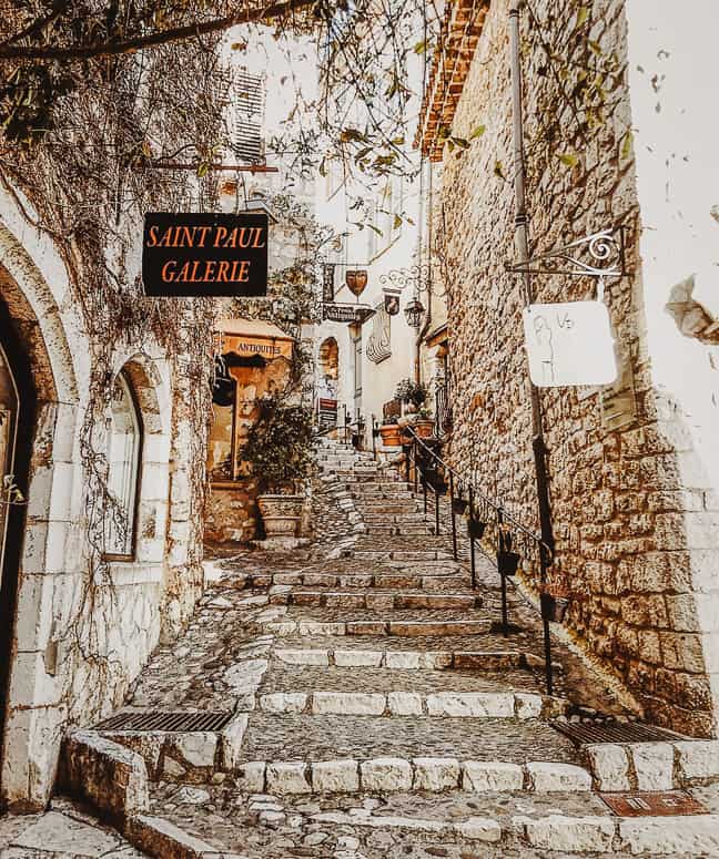 A narrow passageway that is also a staircase in Saint Paul de Vence. the streets and sides of the buildings are cobblestoned, with a tree hanging over the tops of the buildings, and cute signposts for art galleries on display. 