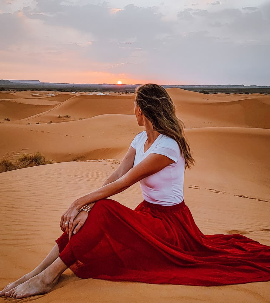 A woman in a white shirt and long red skirt sits on a sand dune in the Sahara, looking behind her innto the distance, as the sun rises on the horizon. 