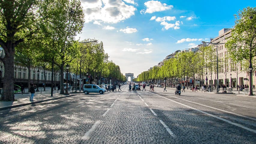 A view down the wide Champs Elysees Avenue. Some people are crossing the street, manicured trees line the road, and the Arc de Triomphe is in the background. 