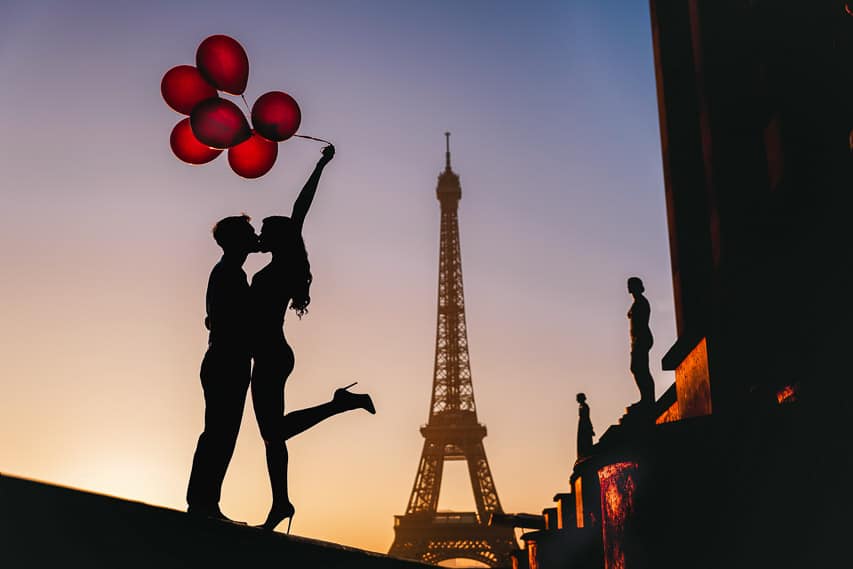 the silhouette of a man and woman kissing in front of the Eiffel Tower, with the woman holding red balloons floating in the wind. An Eiffel Tower photoshoot is a romantic thing to do in Paris for couples. 