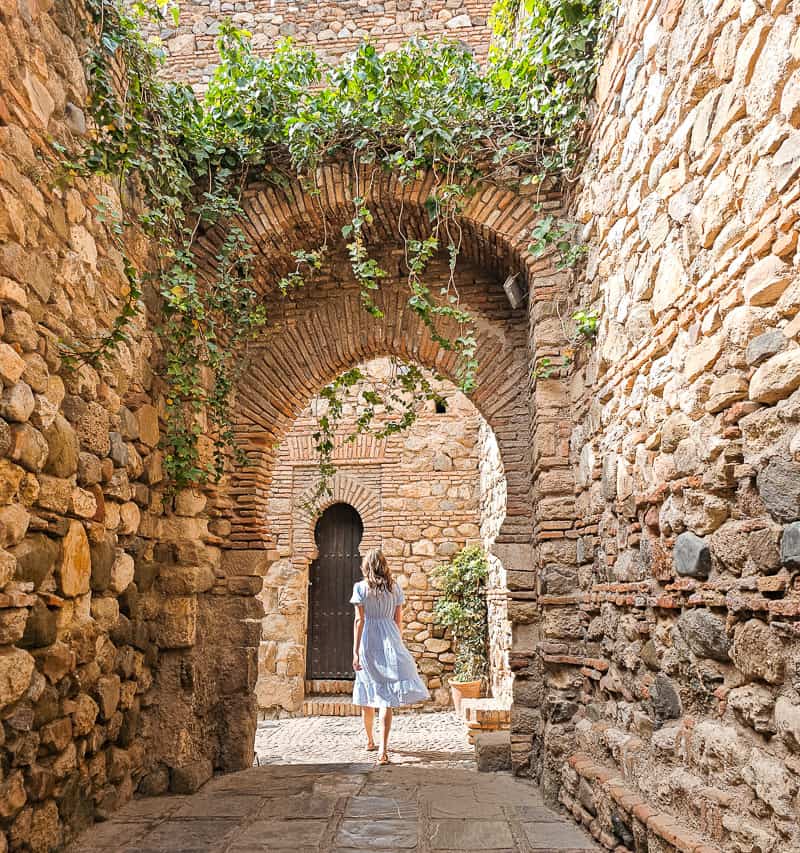 A woman in a blue and white dress walks away from the camera, down a cobblestoned passageway with a large arch at the end and ivy hanging down. The Alcazaba is one of the top reasons to visit Malaga. 