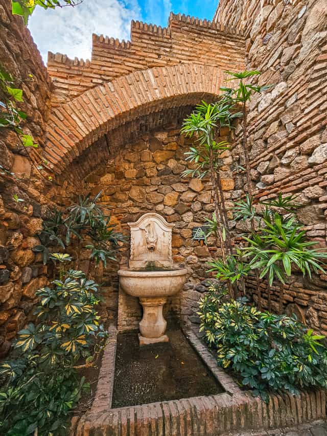 A small corner in the Alcazaba with a small while fountain a curved bricked top, cobblestone walls, and greenery. 