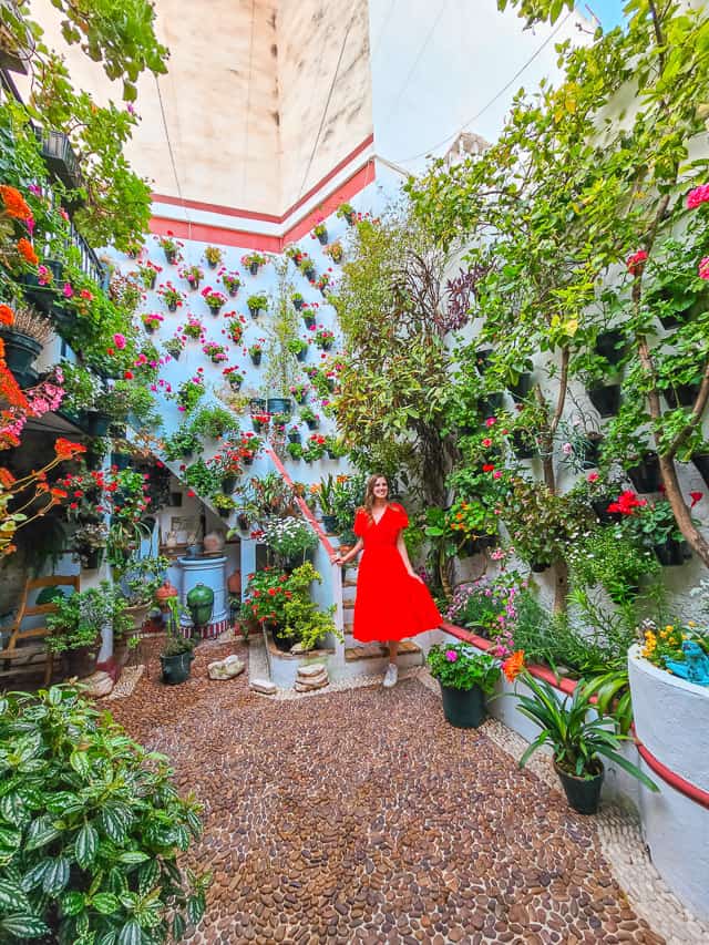 A woman in a red dress walks down a staircase at the back of a courtyard in Cordoba, that has white walls, cobblestoned flooring, plants and trees and flowerpots all over the walls. 