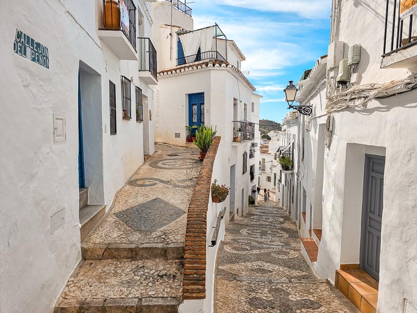 A small street in the village of Frigiliana. The buildings walls are completely white, and the ground has designs in the tilework. 