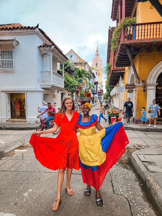 2 women hold the sides of their dresses up together. One of the girls is balancing a bowl of fruit on her head in the streets of Cartagena. 