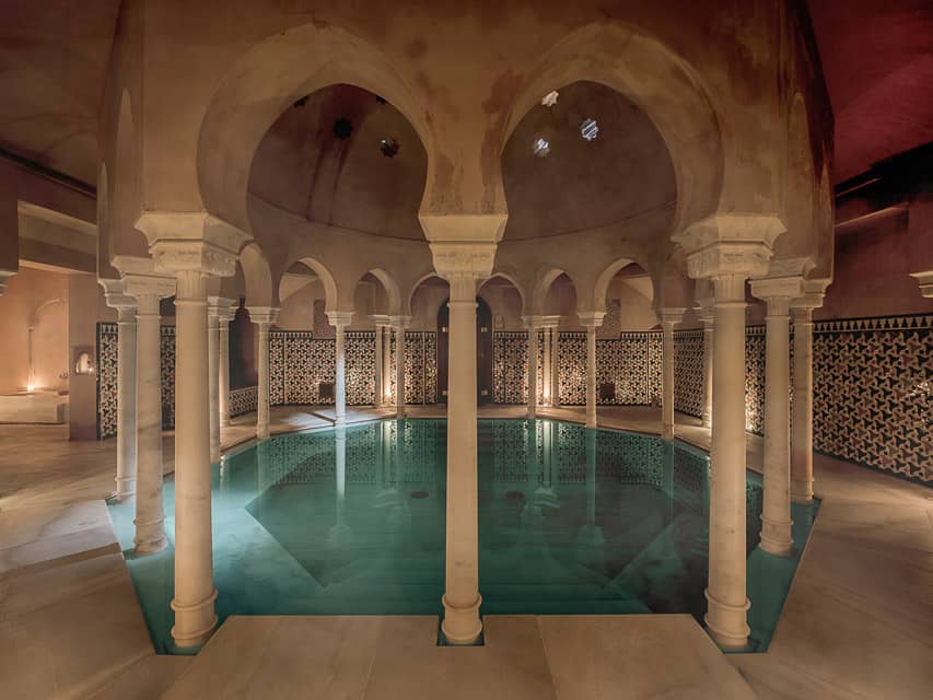 A gorgeous hamman bath with green-blue water in a circular pool, surounded by Arabic archways and columns, and tiles on the walls.