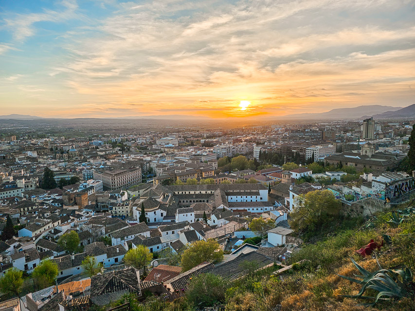 A view from a hill, overlooking the city and the white houses below in the valley, with the sun setting behind some mountains and hills in the distance. 