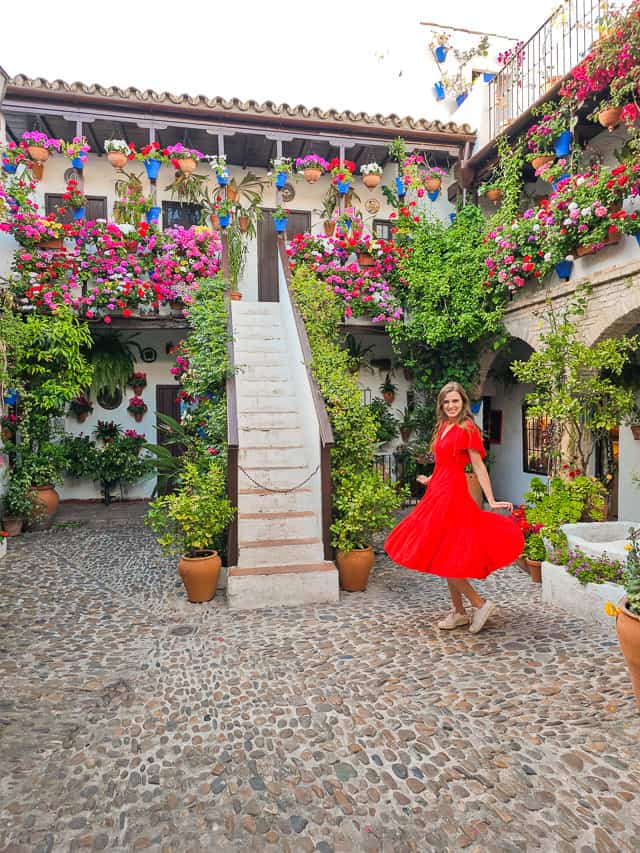 A girl in a red dress twirls in a courtyard full of flowers and greenery. 