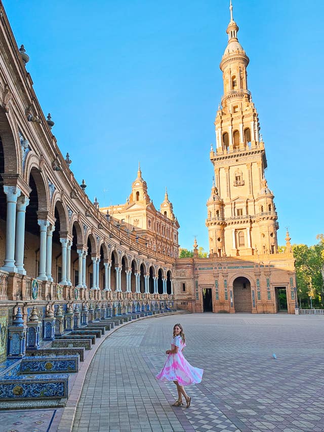 A woman in a pink dress twirls in a beautiful spanish courtyard with the sun just peeking out in the colorful tile courtyard with pillars holding up a castle-like stone building. 