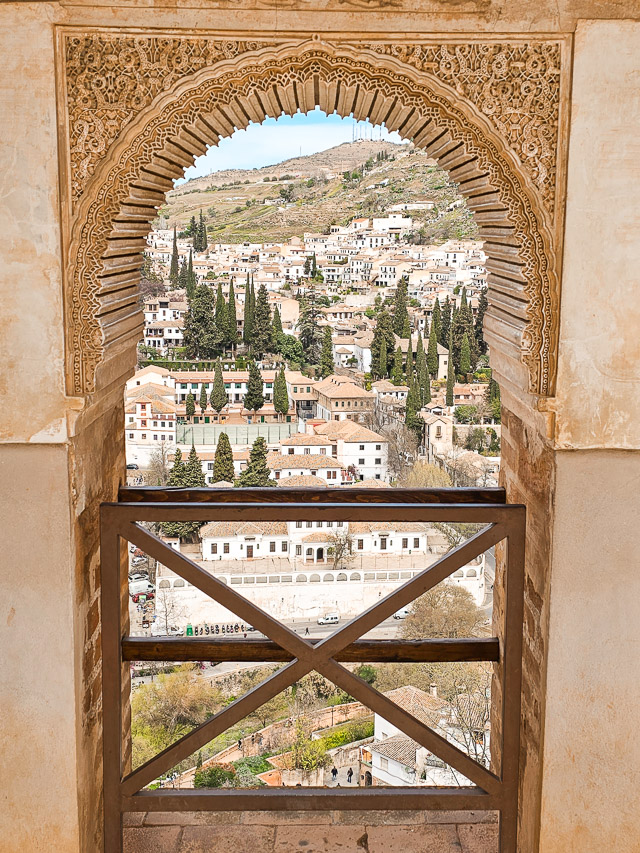A view to the white houses and cypress trees of the Albaicin neighborhood through a fancy window opening in Alhambra. 