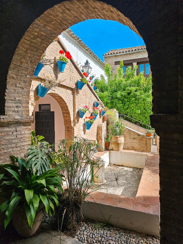 this picture looks through one arch into the corner of a courtyard, where blue flowerpots are hung against bricked steps, and a fountain basin is in front. 