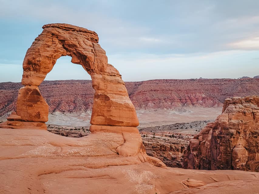 Delicate Arch with orange rocks all around it with reddish rocks behind it.