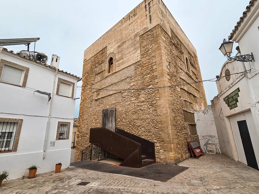 An old stone, square tower sits in the midst of smaller white buildings. The Almohade Tower and Cistern is the remains of an ancient castle.