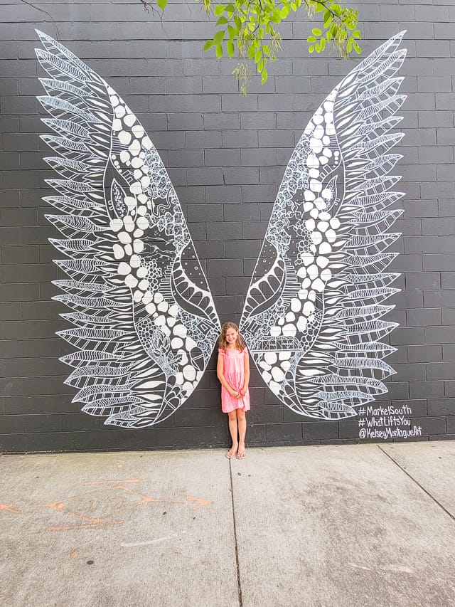 A little girl stands next to a black wall with a large mural of wings.