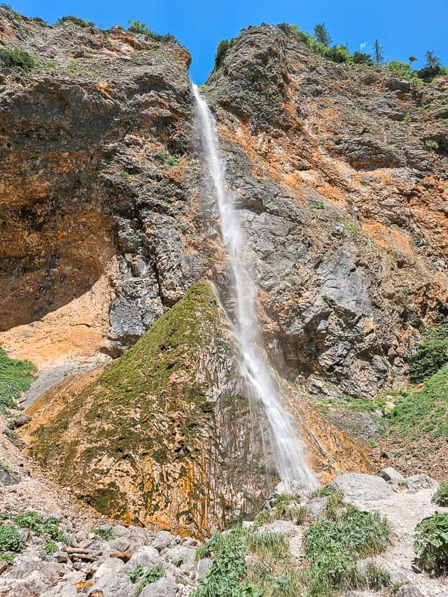 A tall, skinny waterfall goes down over a red cliff.