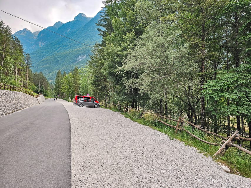 A gravel parking lot on the right side at the trailhead for the Soca River and a concrete street on the left side with trees everywhere and a mountain range in the background. 