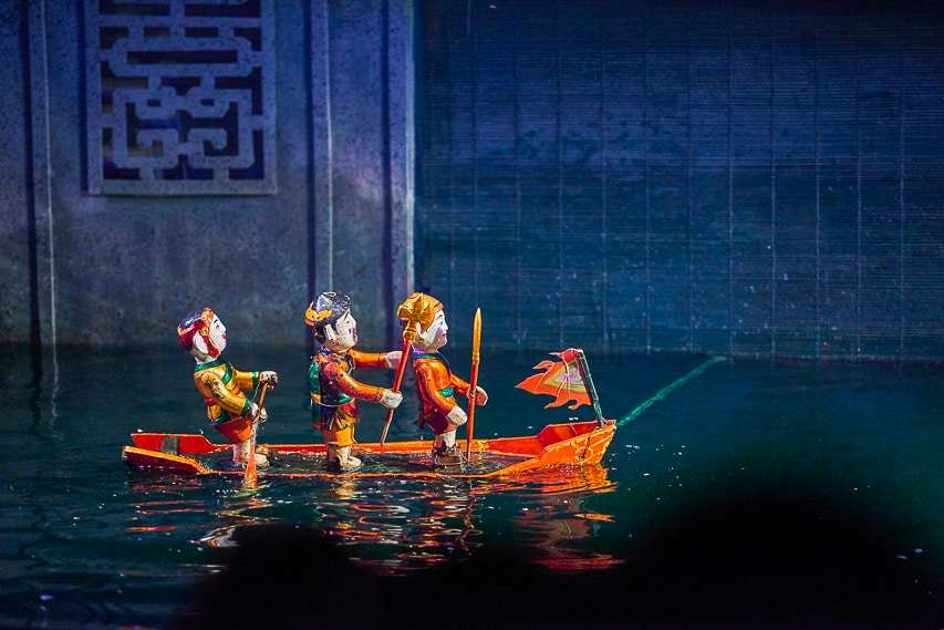 A water puppet show with 3 puppets on a small wood boat in a small tank of water. 
