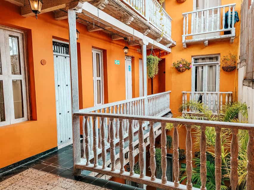 A balcony next to bright orange walls with white wood railings and a palm tree-like plant next to the railing. 