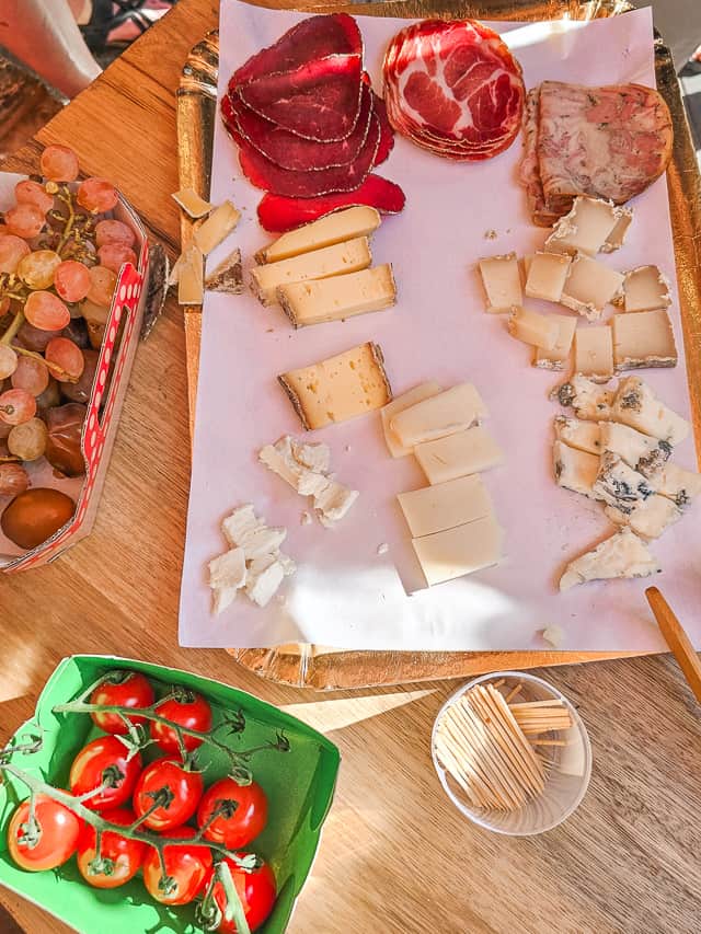 A plate of assorted cheeses with boxes of tomatoes and grapes/dates to the side. At the foot of the cheese board there is a little circular box of toothpicks. 