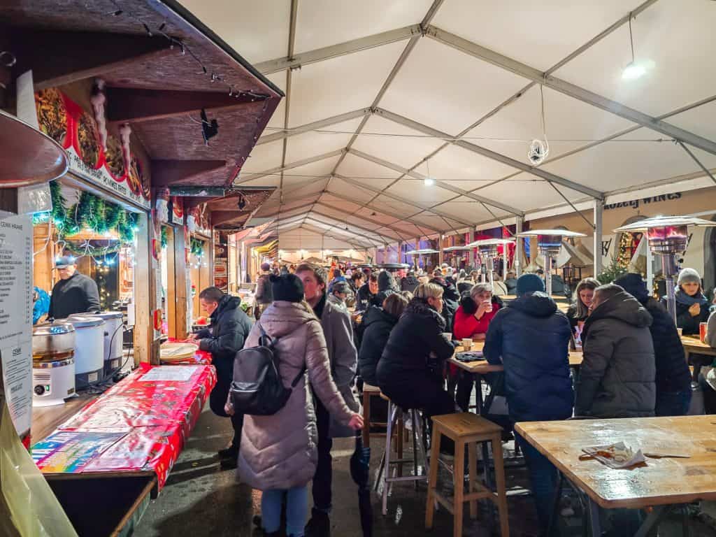 A Christmas market right besides the white cathedral with lots of huts with lights and wood tables. Lots of people in coats are walking around