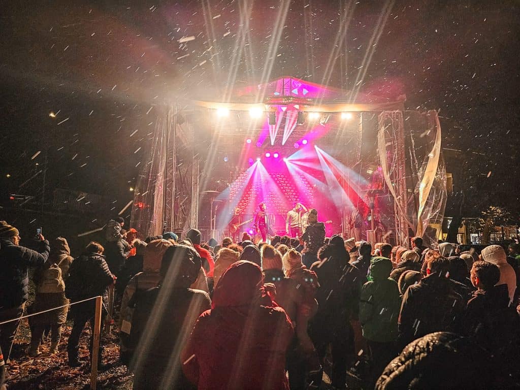 A lit up stage with a band playing music while it was snowing. Lots of people are dancing and singing to the band while crowding around the stage