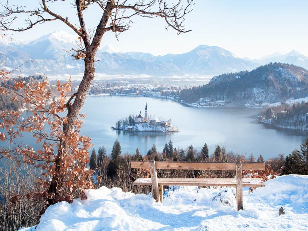 The view of the snow covered Lake bled with an island in the middle. The mountains and city are all dusted with snow. 